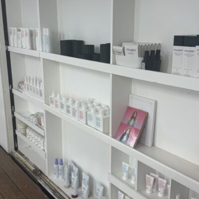 A wide-angle shot of a minimalist wooden salon shelf displaying an organized variety of professional hair care products. The shelf includes various bottles of shampoo, conditioner, styling oils, and tubs of hair balm in neutral-toned packaging. The products are arranged neatly, creating a clean and professional retail display.