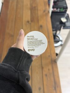 A close-up of a 50g jar of Evo "The Shag" beach paste. The product is in a small, matte white circular tub with minimalist black text. It is a styling product designed for creating messy, matte, beachy texture.