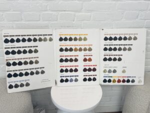 A professional hair color chart spread open on a flat surface, displaying a wide array of synthetic hair swatches. The swatches are organized in rows, ranging from natural blondes and browns to vibrant fashion colors and deep blacks. Each sample is labeled with a specific shade number and name, used by stylists for color consultations.