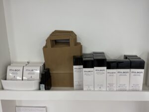 A bright white retail shelf in a salon featuring a collection of Milbon hair care products. The shelf is lined with various professional bottles and tubes, including the Signature series with its distinctive sleek, white and colored packaging. The arrangement is clean and minimalist, showcasing a variety of shampoos, treatments, and styling products.