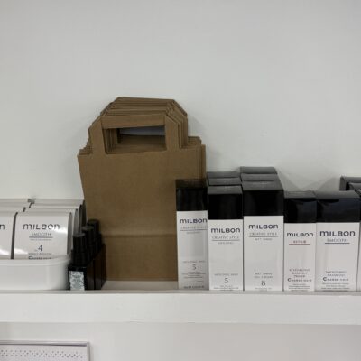 A bright white retail shelf in a salon featuring a collection of Milbon hair care products. The shelf is lined with various professional bottles and tubes, including the Signature series with its distinctive sleek, white and colored packaging. The arrangement is clean and minimalist, showcasing a variety of shampoos, treatments, and styling products.