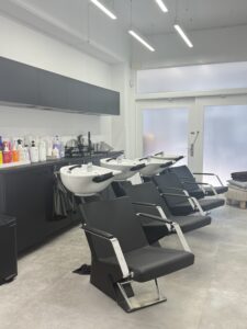 A wide-angle shot of a professional salon’s hair washing station. Four sleek, black ergonomic washbowls with matching reclining chairs are arranged in a clean, uniform row. Four hairstylists are positioned at the stations, draped in professional attire, carefully performing hair treatments and scalp massages for their clients. The room features soft, ambient lighting and a clean, modern aesthetic.