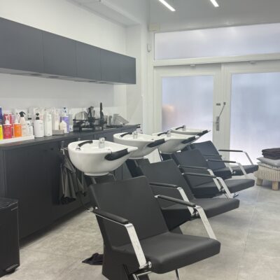 A wide-angle shot of a professional salon’s hair washing station. Four sleek, black ergonomic washbowls with matching reclining chairs are arranged in a clean, uniform row. Four hairstylists are positioned at the stations, draped in professional attire, carefully performing hair treatments and scalp massages for their clients. The room features soft, ambient lighting and a clean, modern aesthetic.