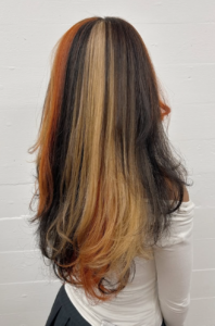 A back view of the previous style, showing a textured blend of black, vibrant orange, and blonde hair. The colors are distributed in a chunky, layered pattern that creates a high-energy, multi-colored look from behind.