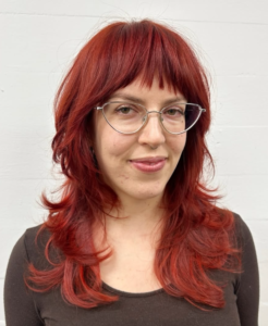 A front profile portrait of a deep, warm red-orange or burnt sienna hair color. The tone leans heavily into red tones, and the hair is styled with soft texture around the face.
