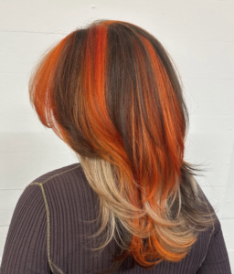 A back view of a high-contrast calico color style. The hair is a patchwork blend of bright orange, deep chocolate brown, and sandy blonde sections, creating a dynamic, multi-colored pattern throughout the lengths.
