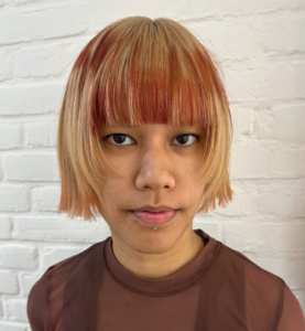 A front profile of a chin-length platinum blonde bob with full, straight-across bangs. The fringe features a creative, fragmented design of bright orange color applied in horizontal, blocky strokes to create a visual "glitch" effect against the blonde hair.