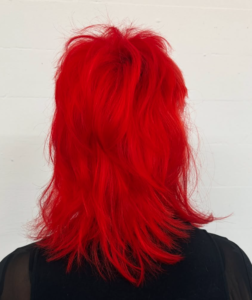A back profile view of a textured, short-to-medium length mullet dyed in a vibrant, neon red. The hair features choppy, spiky layers on top and at the crown, with the back length falling just to the shoulders. The color is a bold, high-saturation crimson that stands out against the skin.