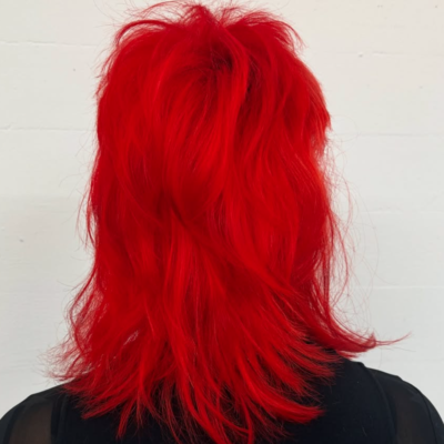 A back profile view of a textured, short-to-medium length mullet dyed in a vibrant, neon red. The hair features choppy, spiky layers on top and at the crown, with the back length falling just to the shoulders. The color is a bold, high-saturation crimson that stands out against the skin.