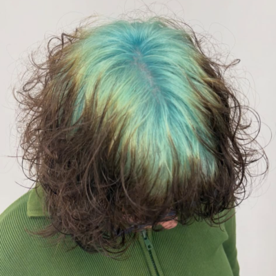 A front view of a short bixie haircut where the client is looking down, causing the top layer of natural brown hair to cascade slightly forward. This position reveals a prominent, contrasting band of vibrant teal-mint green color applied directly at the roots across the crown and parting line, characteristic of the "ghost root" technique.