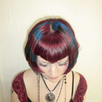 A high-angle front profile showcasing a dramatic creative transformation. The hair is dyed a vivid, all-over crimson red with a sharp V-shaped fringe (v-bangs) at the forehead. The upper angle clearly highlights the precision of the V-cut and the intense saturation of the red pigment at the crown.