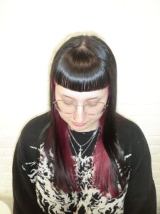 A front profile taken from an upper angle, looking down at a high-contrast peekaboo hair transformation. The top layer of the hair is a deep jet-black, featuring full, straight-across bangs. From underneath, a vibrant, saturated bright red color is visible, creating a bold two-tone effect that peeks through the black layers and frames the face.