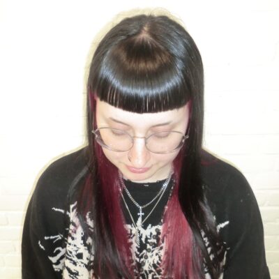 A front profile taken from an upper angle, looking down at a high-contrast peekaboo hair transformation. The top layer of the hair is a deep jet-black, featuring full, straight-across bangs. From underneath, a vibrant, saturated bright red color is visible, creating a bold two-tone effect that peeks through the black layers and frames the face.
