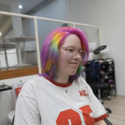 A side profile view of the hair dyed a vibrant, all-over pink magenta. Beneath the top layer, a hidden peekaboo section features horizontal stripes of red, orange, yellow, green, blue, and purple to create a vivid rainbow effect. A single distinct strand on the surface is also patterned with these same horizontal rainbow stripes, creating a sharp, colorful contrast against the solid magenta base.
