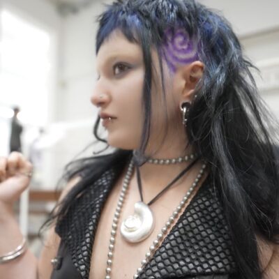 A front profile showcasing an edgy hairstyle with deep blue and purple tones concentrated at the roots. The sides of the head are closely shaved, featuring an intricate, clean spiral graphic design etched directly into the undercut, while the colorful, longer top sections are styled to reveal the shaved hair art.