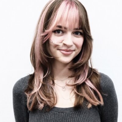 A front profile showing a bold hair transformation. The hair has been cut to chest-length and styled straight. It features large, chunky pink highlights that contrast sharply against a dark brunette base. The pink sections are particularly prominent around the face-framing layers and crown, creating a vibrant, color-blocked aesthetic.