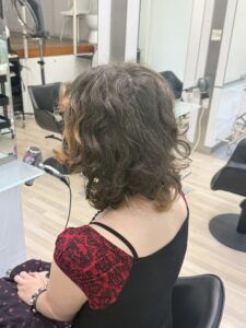 A back view of shoulder-length medium brown hair. The color is mostly solid with some faded, unevenly bleached parts toward the ends. The hair has a natural curly texture and a simple, blunt hemline.