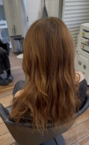 A back view of long, naturally wavy hair in a rich chocolate brown shade. The waves are loose and textured, cascading down the back with a soft, voluminous finish. The color is consistent and warm, showing off the hair’s natural shine.