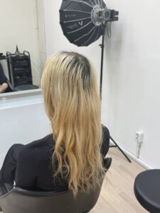 A back view of long, straight hair featuring a pale blonde canvas. There is a visible natural dark root grow-out of a few inches, transitioning into a light, platinum blonde through the mid-lengths and ends.