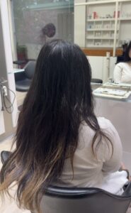 A back view of long, straight black hair. The top and mid-lengths are a solid jet-black, transitioning into uneven, faded blonde bleached ends. The hair has a natural texture with some visible dryness at the lightened tips.