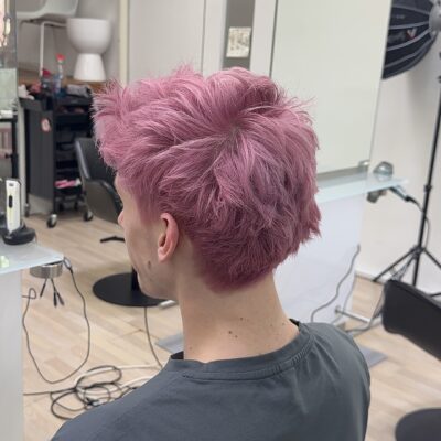A back profile of the same man after a bold color transformation. His hair is now a vibrant, cool-toned orchid pink with subtle lavender undertones. The hair maintains its textured, voluminous style, with the bright pink hue reaching from the roots to the tips, creating a modern and edgy look.
