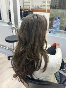 A back view of long, naturally wavy hair featuring a blend of medium brown and golden blonde tones. The hair cascades down the back in soft, beachy waves with a textured finish, showcasing a lived-in, sun-kissed color palette.