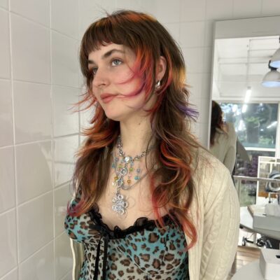 A front profile of a vibrant "sunset" hair transformation. The long, wavy brown-blonde hair features a "halo" of color around the face and crown, blending shades of soft purple, vivid pink, and warm orange into the blonde. The same sunset gradient is repeated at the ends in a dip-dye style, creating a colorful frame of pink and orange against the natural lengths.