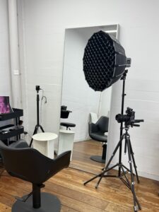 A behind-the-scenes view of a professional salon filming setup. A client is seated in a black styling chair in front of a large, well-lit mirror. Positioned in front of them is a tripod-mounted smartphone with a ring light, capturing a high-definition video of the hair transformation process. The background shows a clean, modern salon environment with styling tools and products neatly arranged.