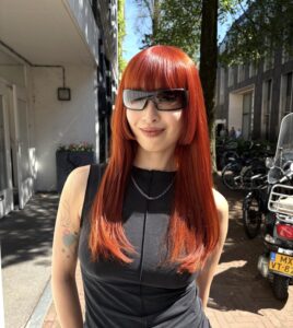 A front profile showcasing a dramatic transformation to a vivid, bright orange hair color. The hair has been reshaped into a structural Hime cut, featuring a thick, straight-across fringe and blunt, cheek-length side-locks that frame the face. The remaining lengths are kept long and straight, highlighting the intense saturation of the copper-orange hue and the sharp geometric lines of the cut.