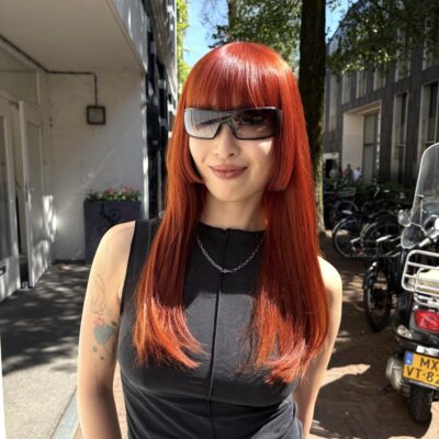 A front profile showcasing a dramatic transformation to a vivid, bright orange hair color. The hair has been reshaped into a structural Hime cut, featuring a thick, straight-across fringe and blunt, cheek-length side-locks that frame the face. The remaining lengths are kept long and straight, highlighting the intense saturation of the copper-orange hue and the sharp geometric lines of the cut.