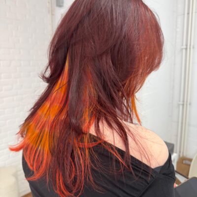 A back view of the vibrant red hairstyle. The top layer is a solid, intense red, which lifts to reveal a hidden "peekaboo" underlayer of bright neon orange. The sides of the hair show the continuation of the "raccoon tail" striped pattern, creating a high-contrast, multi-dimensional look that pops against the dark red surface.