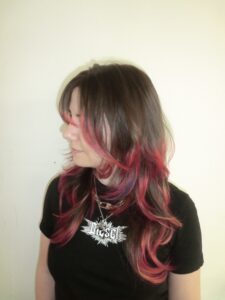A side profile showcasing a vibrant "flame effect" dip-dye on a fresh layered cut. The natural brown hair transitions dramatically near the mid-lengths into a vivid, multi-tonal gradient of deep purple, fiery red, and bright neon orange at the tips. The texturized, face-framing layers emphasize the movement of the colors, making the ends look like flickering flames.