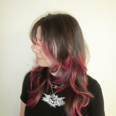 A side profile showcasing a vibrant "flame effect" dip-dye on a fresh layered cut. The natural brown hair transitions dramatically near the mid-lengths into a vivid, multi-tonal gradient of deep purple, fiery red, and bright neon orange at the tips. The texturized, face-framing layers emphasize the movement of the colors, making the ends look like flickering flames.